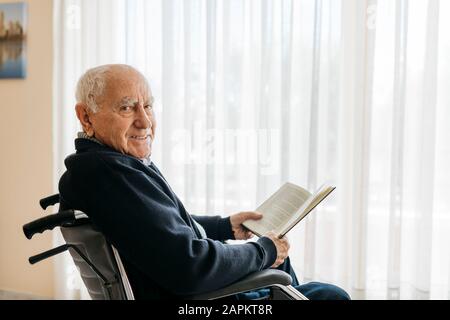 Porträt eines lächelnden älteren Mannes, der mit einem Buch im Rollstuhl sitzt Stockfoto