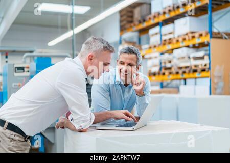 Zwei lächelnde Kollegen, die Laptop benutzen und in einer Fabrik diskutieren Stockfoto