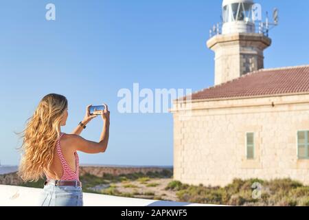 Junge Frau fotografiert den Leuchtturm von Punta Nati auf Menorca, Spanien Stockfoto