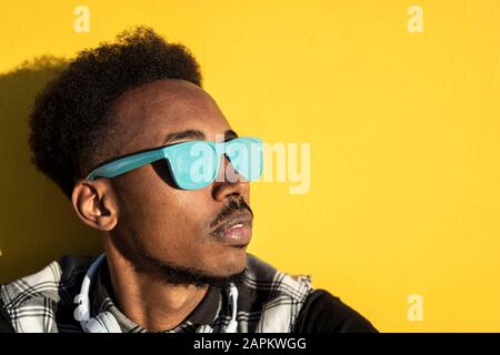 Porträt des jungen Mannes, der eine hellblaue Sonnenbrille gegen die gelbe Wand trägt Stockfoto