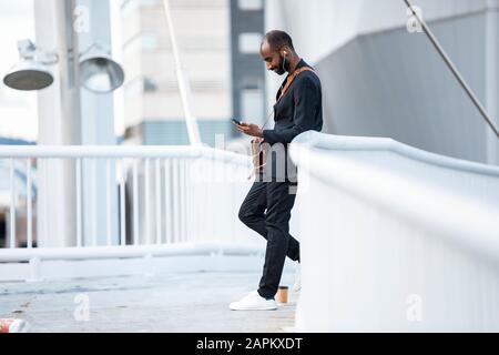 Junger Geschäftsmann mit Ohrhörern, die auf einer Fußgängerbrücke stehen und auf das Smartphone blicken Stockfoto