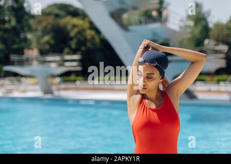 Porträt der jungen Frau mit Schwimmkappe und Schutzbrille in rotem Badeanzug vor dem Pool Stockfoto