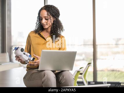 Junge Geschäftsfrau sitzt mit Laptop und Roboter auf dem Schreibtisch im Büro Stockfoto