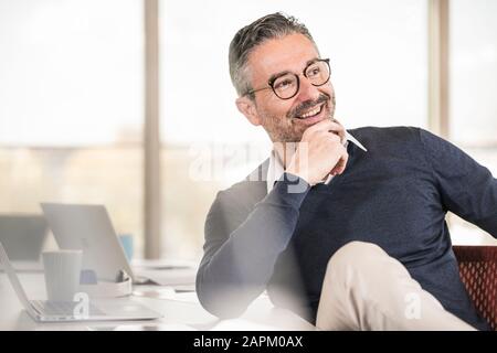 Lächelnder reifer Geschäftsmann, der am Schreibtisch im Büro sitzt Stockfoto