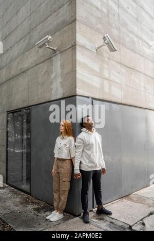 Mann und Frau lehnen sich an eine Wand, Sicherheitskameras hängen über ihren Köpfen Stockfoto