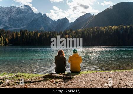 Rückansicht des Paares, das am See in Laghi di Fusine, Friuli Julisch Venetien, Italien sitzt Stockfoto