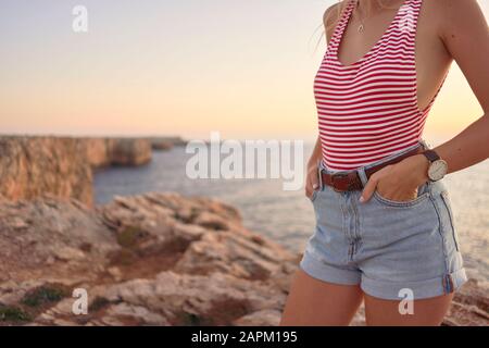 Junge Frau, die Sonnenuntergang am Meer genießt und auf einer Klippe steht Stockfoto
