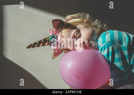 Porträt des kleinen blonden Mädchens, das ein Einhorn trägt, das sich an den rosafarbenen Ballon lehnt Stockfoto