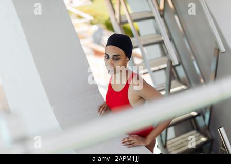 Porträt der jungen Frau mit Schwimmkappe und rotem Badeanzug, der auf einer Treppe auf dem Hochbrett steht Stockfoto