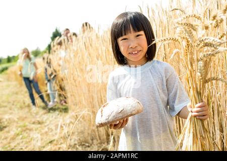 Kleines asiatisches Mädchen, das auf dem Feld steht und Brotlaib und Weizenähren hält Stockfoto