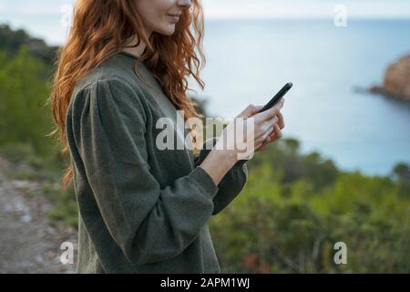 Redheaded junge Frau mit Handy an der Küste, Ibiza, Spanien Stockfoto