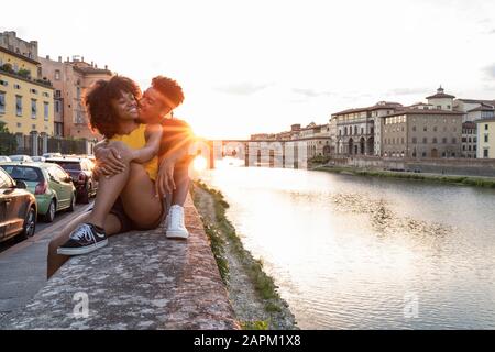 Liebevolles junges Touristenpaar, das bei Sonnenuntergang an einer Wand am Arno sitzt, Florenz, Italien Stockfoto