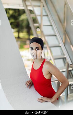 Porträt der jungen Frau mit Schwimmkappe und rotem Badeanzug, der auf einer Treppe auf dem Hochbrett steht Stockfoto