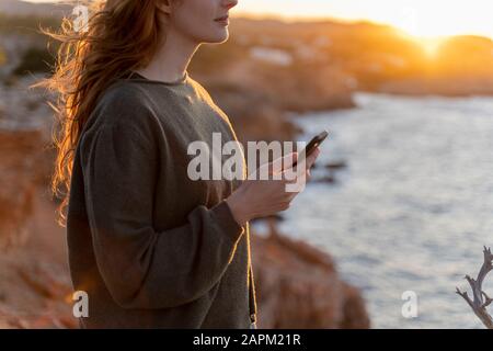 Redheaded junge Frau mit Handy an der Küste bei Sonnenuntergang, Ibiza, Spanien Stockfoto