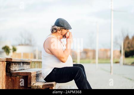 Mann mit Bierbauch, der auf Stufen sitzt und ein Sandwich isst Stockfoto