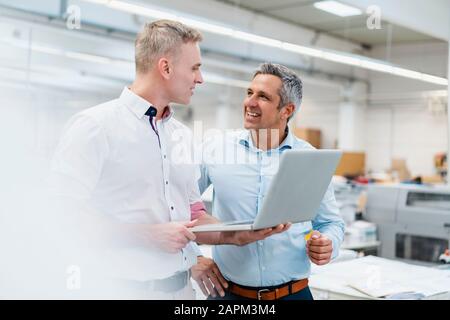 Zwei Kollegen, die Laptop verwenden und in einer Fabrik diskutieren Stockfoto