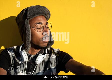 Porträt des jungen Mannes, der mit Kopfhörern mit Kappe und einer metallumrandeten Brille gegen die gelbe Wand getragen wird Stockfoto