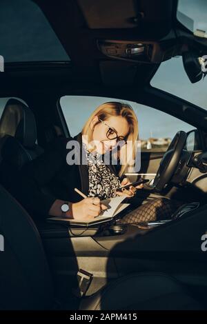 Junge blonde Frau, die Smartphone im Auto benutzt und in einem Notizbuch schreibt Stockfoto