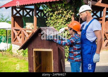 Junge, Teenager, der mit dem Schutzhelm beschäftigt ist, der sich überstellt, das Hundehaus mit seinem Vater zu reparieren. Vater lehrt kleinen Sohn, mit einem Bandli richtig zu messen Stockfoto