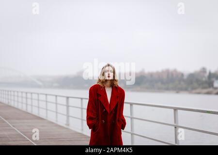 Porträt der jungen Frau, die an regnerischen Tagen roten Mantel auf einer Brücke trägt Stockfoto