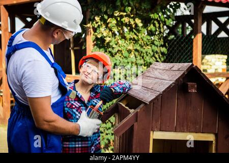 Vater und sein Sohn arbeiten in einer Holzwerkstatt zusammen. Sie konzentrieren sich auf den Bau eines Holzhundehauses, der Sohn sitzt darin mit einem Lack b Stockfoto