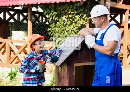 Vater und sein Sohn arbeiten in einer Holzwerkstatt zusammen. Sie konzentrieren sich auf den Bau eines Holzhundehauses, der Sohn sitzt darin mit einem Lack b Stockfoto