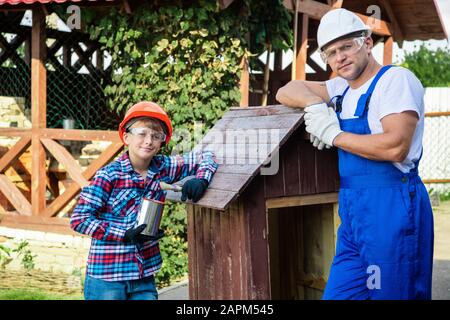 Vater und sein Sohn arbeiten in einer Holzwerkstatt zusammen. Sie konzentrieren sich auf den Bau eines Holzhundehauses, der Sohn sitzt darin mit einem Lack b Stockfoto