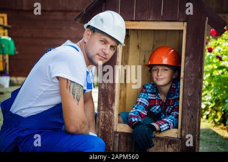 Vater und sein Sohn arbeiten in einer Holzwerkstatt zusammen. Sie konzentrieren sich auf den Bau eines Holzhundehauses, der Sohn sitzt darin mit einem Lack b Stockfoto