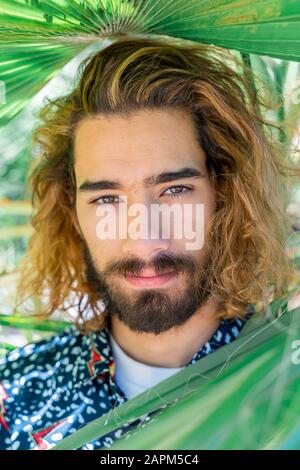 Porträt eines bärtigen jungen Mannes mit gefärbten Haaren unter den Palmblättern Stockfoto