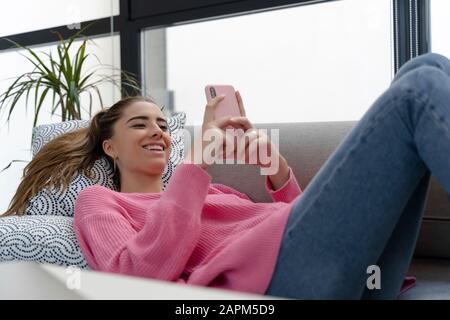 Porträt einer glücklichen jungen Frau, die mit dem Handy auf dem Sofa liegt Stockfoto