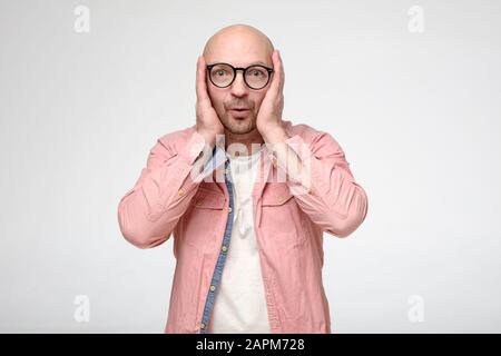 Zufriedener Mann hält emotional die Hände im Gesicht, er freut sich über eine unerwartete, angenehme Überraschung und schaut gerne auf die Kamera. Stockfoto
