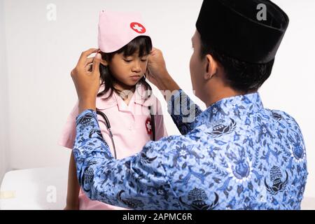 Asiatischer Vater, der Batik korPRI trägt, kleidet seine Tochter in einer Arztuniform, bevor er zur Schule geht Stockfoto