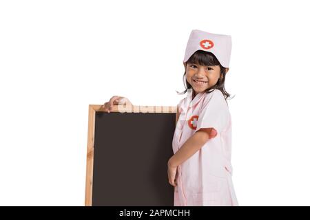 Asiatisches kleines Mädchen, das eine Krankenschwester Uniform mit Hand trägt, die eine Tafel neben ihr auf weißem Hintergrund hält Stockfoto