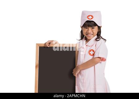 Lächelnde kleine Krankenschwester, die eine Uniform trägt und eine Tafel neben ihr auf einem isolierten Hintergrund hält Stockfoto