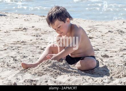 Ein kleiner Junge spielt glücklich an einem sonnigen Strand und baut während seines Familienurlaubs ein Sandschloss Stockfoto