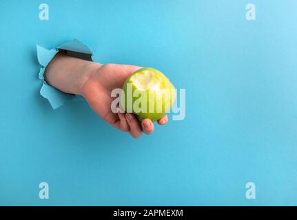 Die Hand einer Frau hält einen gebissenen apfel durch ein Loch im Papier auf blauem Grund. Kopierbereich Stockfoto