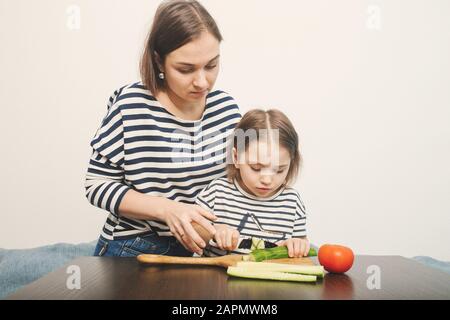 Die junge Mutter lehrt ihre kleine Tochter, Gemüse für gesunden Salat zu schneiden. Weißer Hintergrund. Echte Menschen zu Hause. Stockfoto