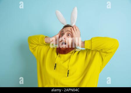 Schockiert, staunend. Kaukasischer Mann als Osterhase mit heller lässiger Kleidung auf blauem Studiohintergrund. Frohe ostergrüße. Konzept menschlicher Emotionen, Gesichtsausdruck, Feiertage. Copyspace. Stockfoto