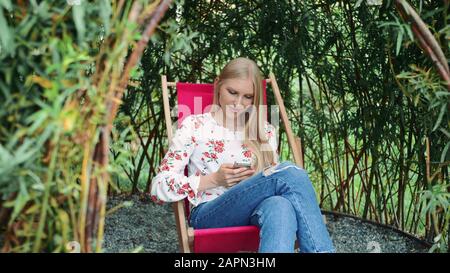 Junge Frau mit Smartphone in Pflanzengazebo. Schöne junge Dame lächelt und tippt auf dem Smartphone, während sie in einem fantastischen Pflanzengazebo im Park sitzt. Stockfoto
