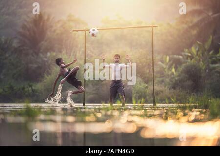 Der Junge, der einen Fußballball trickst, die Gruppe der Kinder, die in Thailand einen Fußball auf dem Naturfluss spielen. Stockfoto