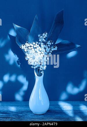 Blumenstrauß aus Liliensteinen des Tals in einer weißen Vase mit sonnigen Höhepunkten in klassischer blauer Farbe. Grußkarte. Sommermorgen. Stockfoto