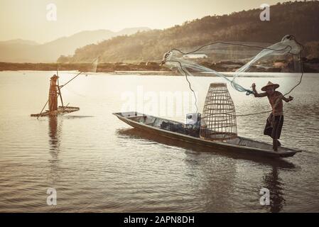 Thai Fischer auf Holzboot wirft ein Netz für den Fang von Süßwasserfischen in Natur Fluss am frühen Abend vor Sonnenuntergang Stockfoto