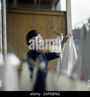 Frau hängt die frisch gewaschene Kleidung, die an der Wäscheleine trocknet, mit einem Hängefach mit Wäscheklappen, 1965. Stockfoto