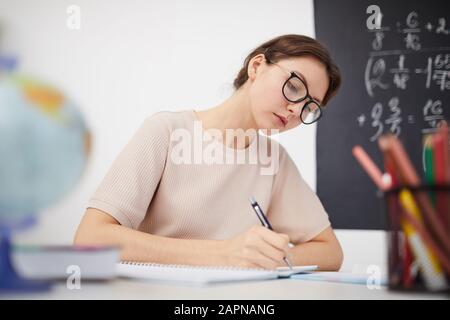 Junge Lehrerin in Brillen, die am Tisch sitzen und in der Schule im Unterricht Papierarbeit leisten Stockfoto