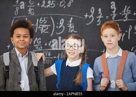 Porträt der Gruppe von Schulkindern, die mit Rucksäcken stehen und im Hintergrund die Kamera vor der Tafel betrachten Stockfoto