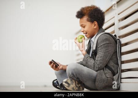 Afrikanischer Schuljunge, der auf dem Boden sitzt und Apple isst und etwas auf dem Handy beobachtet, ruht er nach dem Unterricht Stockfoto