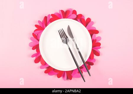 Runde weiße Platte mit schwarzer Gabel und einem Messer auf einem rosafarbenen Tisch mit Papierherzen. Valentinstag Konzept und legte Tisch. Stockfoto