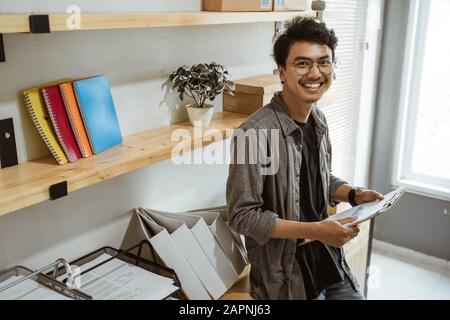 Pose des lächelnden jungen asiatischen Unternehmers, wenn er eine Zwischenablage an der Raumarbeit hält Stockfoto