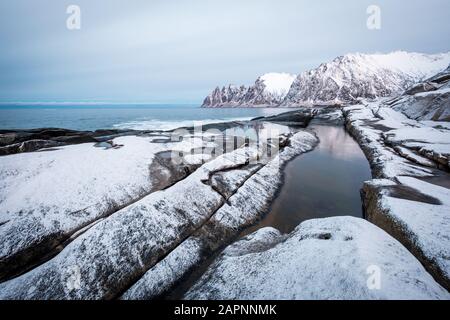 Blick über den Ersfjord von Felsen und Rockpools, die mit Neuschnee bedeckt sind, zu den Bergen von Oksen an einem dunklen bewölkten Tag im Winter, Kap Tungeneset Senja, Norwegen Stockfoto