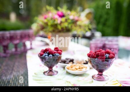 Schönen Gläser von Violett Glas auf dem Tisch mit Beeren und Blumen, Komposition für Catering im Freien Stockfoto